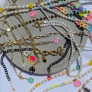 A bunch of colourful beaded necklaces tangled together against a grey slate background. Necklaces made of polymer clay beads, freshwater pearl beads, acrylic themed beads and 18K gold-plated details. 